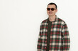 © Oleksandr Zinchenko - A man stands confidently wearing sunglasses and a plaid shirt. He smiles while posing against a plain white wall, creating a relaxed and casual atmosphere