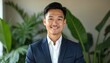 © miss irine - Asian man in suit smiles. Confident business professional in corporate office. Green plants, fresh background. Presentation profile photo with charisma. Young modern guy in business setting.