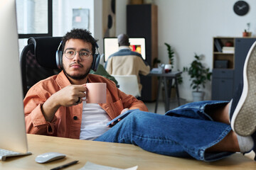Poster - Young restful male office worker in headphones having coffee at break and looking at computer screen while watching tutorial