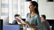 © fizkes - Positive young company leader woman holding digital tablet, standing in office space with colleagues working behind, looking away, smiling, thinking on business plan, Internet marketing. Banner shot