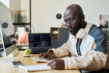 Fototapete - Young serious African American man in casual attire sitting by workplace with desktop computer and taking notes in notebook