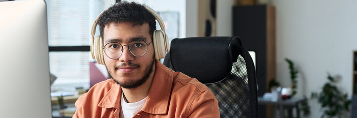Poster - Header of young smiling man in eyeglasses and headphones looking at camera during network while sitting in office