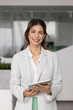 © fizkes - Happy successful Hispanic executive woman holding digital tablet, posing in office, looking at camera, smiling. Latin female business leader, company owner vertical portrait