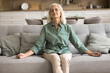 © fizkes - Mindfulness and meditation. Calm peaceful older female pensioner rest on couch at home practice easy yoga exercises hold fingers in mudra signs breath deep with closed eyes meditate try to reach zen