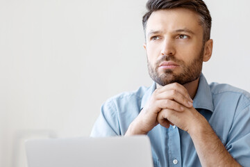 Poster - Portrait Of Pensive Millennial Businessman Sitting At Workplace With Laptop And Looking Away, Serious Male Entrepreneur Thinking About Business Ideas, Resting Chin On Hands, Closeup With Copy Space