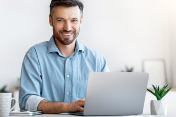 Poster - Smiling Young Businessman Sitting At Desk With Laptop In Modern Office, Looking At Camera, Free Space