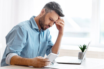 Poster - Worried male freelancer looking at smartphone screen while working at home office, received bad news, got scam message, thoughtful man touching forehead with concerned face expression, copy space