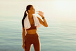© Prostock-studio - Wellness Concept. Active girl drinking mineral water from bottle, standing on the beach in wireless earphones