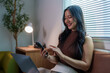 © crizzystudio - Young asian woman working from home, sitting on a comfortable sofa and using smartphone and laptop, illuminated by the warm light of a desk lamp