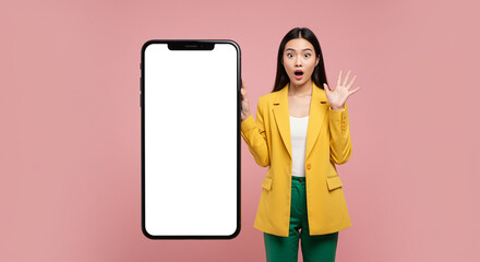 Poster - Smartphone mockup held by surprised Asian woman in yellow jacket presenting blank screen for mobile app promotion or advertising. Studio shot of excited girl showing phone display for website design
