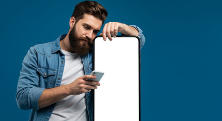 Poster - Bearded man in denim shirt leans on large smartphone while using smaller smartphone against blue background. Man holds smartphone in hand and looks at its screen