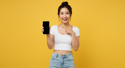Poster - Woman holding smartphone pointing finger with happy expression against yellow background. Asian woman showing mobile phone screen with smile in studio shot