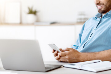 Poster - Handsome young businessman listening music on smartphone with headphones while working at office, sitting at workplace with laptop, browsing internet or messaging on cellphone, free space