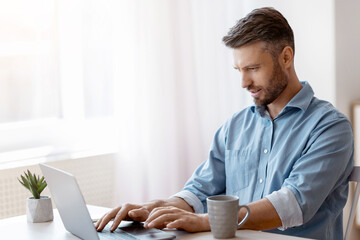 Poster - Unemployed man scrolling job search website at home using laptop computer, handsome male freelancer working remotely at desk near window, typing on keyboard, answering emails, copy space