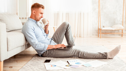 Poster - Home Workplace. Side view of young serious man working in living room, sitting on the floor with laptop, drinking coffee. Focused professional guy using computer, reading business news