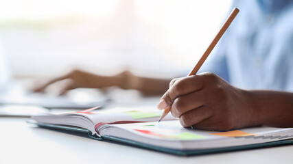 Poster - Unrecognizable african female secretary taking notes while working on laptop in office, writing in notepad to-do list, planning business schedule, selective focus on arm with pencil, closeup