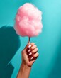 © Will - A hand holding a stick with cotton candy, neatly manicured nails painted in a light pink color, matching the cotton candy. The bright lighting casts a hard shadow on the pastel blue background.