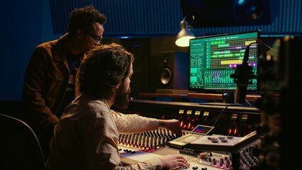 Fototapete - Team of audio technician and artist producing and editing sounds on mixer console, using professional software on pc. Musician does collaboration with engineer to create music. Camera B.