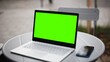 © VAKSMANV - View of Caucasian woman sitting at table in front of laptop with green screen. In meantime using smartphone with chroma key on display. Scrolling with one finger on phone. Sipping coffee.