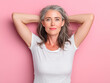 © Helena GARCIA - Mujer de mediana edad con pelo canoso, vistiendo camiseta blanca, apoyando sus brazos en la cabeza feliz, sobre fondo rosa pastel
