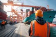 © Hotpin - Dock worker in orange safety vest and helmet observing cargo operations at busy shipping port with cranes, containers, and freight ship on a clear sunny day