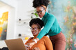 © Studio Marmellata - A woman in a green sweater leans over to help her colleague with a laptop in a bright office with modern decor, featuring indoor plants and a clothing rack.
