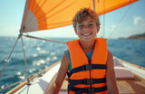 Young boy smiles, wearing orange life vest, sailing yacht on blue sea. Kid enjoys water sport, active summer vacation on sunny day. Happy child enjoys boating with sailboat, ocean travel adventure.