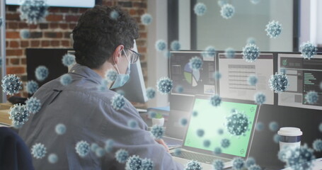 Poster - Image of covid 19 cells floating over man wearing face mask, using laptop in office