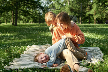 Wall Mural - Candid moment of mother with two children playing and laughing together while sitting on picnic blanket in park copy space