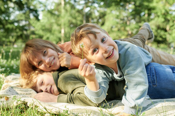 Wall Mural - Portrait of cheerful young boy holding flower and smiling at camera while playing with family on picnic blanket in sunlit park