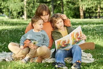 Wall Mural - Full length portrait of caring mother with two boys sitting on blanket and reading storybook enjoying time together outdoors in green park