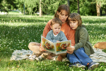 Wall Mural - Full length scene of mother with two children sitting on picnic blanket and reading book enjoying time together outdoors in green park