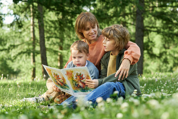 Wall Mural - Candid scene of mother and sons reading book sitting together on picnic blanket in green Summer park copy space