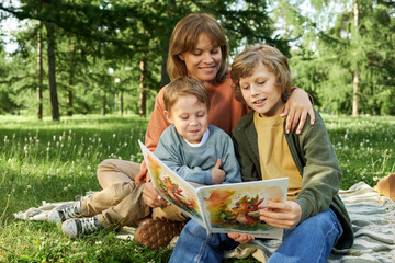 Wall Mural - Portrait of mother and two children reading book together outdoors on picnic blanket in green Summer park copy space