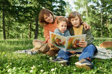 Wall Mural - Full length portrait of smiling woman with two children reading book together sitting on blanket outdoors in green Summer park copy space