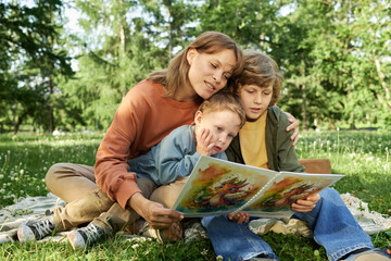 Wall Mural - Portrait of caring young woman as mother reading book to two children while sitting together on green grass in sunny Summer park