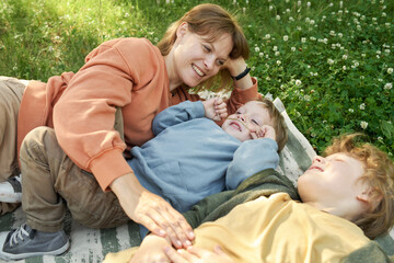 Wall Mural - Candid scene of happy family mother and two boys lying on picnic blanket together and having fun smiling joyfully 