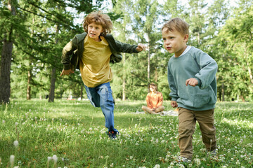 Wall Mural - Full length portrait of two young brothers running on green grass in park with mother watching in background