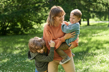 Wall Mural - Candid scene of happy mother playing with sons outdoors in green sunlight meadow and holding child