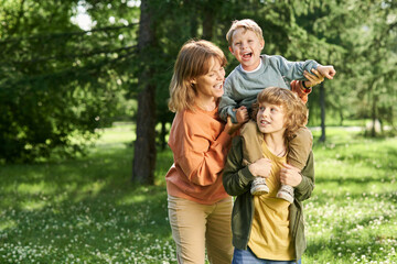 Wall Mural - Waist up portrait of young boy holding little brother on shoulders with mother helping family enjoying time together in green park copy space 