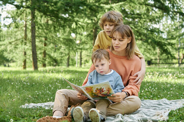 Wall Mural - Full length portrait of mother with two boys reading book together outdoors sitting on picnic blanket in sunlit green park
