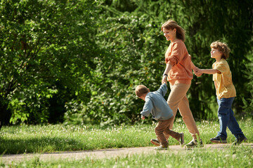 Wall Mural - Full length side view of carefree mother with two boys holding hands and walking on path in green forest together lit by sunlight copy space