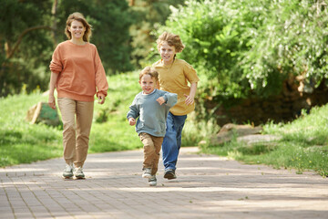 Wall Mural - Full length front view of two brothers running towards camera on path in green park with mother walking in background copy space
