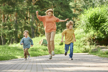 Wall Mural - Full length front view of cheerful mother with two boys running playfully towards camera in green park copy space