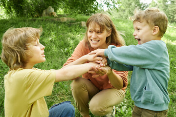 Wall Mural - Candid scene of smiling mother playing with two sons in park family stacking hands together in sunny outdoor setting