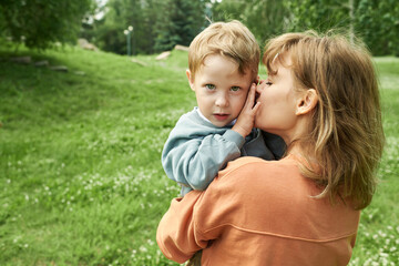 Wall Mural - Back view of mother holding son outdoors in green park and kissing cheek with young boy looking at camera copy space