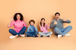 © deagreez - Happy family of four sitting together on a beige background while making playful hand gestures, showcasing love and bonding