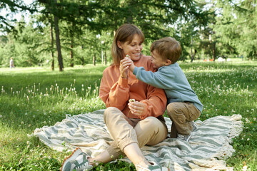 Wall Mural - Full length scene with young boy giving flower to smiling mother while having fun in green park together sitting on picnic blanket