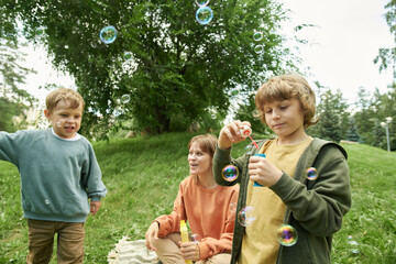 Wall Mural - Portrait of young boy blowing bubbles in park outdoors with family relaxing on picnic blanket in background, copy space