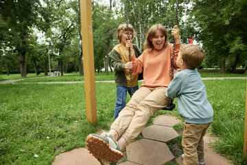 Wall Mural - Portrait of happy family with two children at playground in park, with two boys pushing mom on swing and having fun together